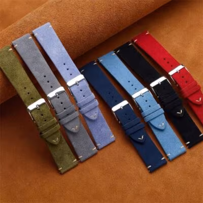 Seven colorful suede replacement watch bands lined up side-by-side. Colors featured are olive green, grey, light blue, navy blue, mid-blue, black, and red.
