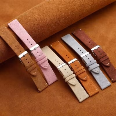 Six suede watch straps in soft earthy and pastel tones, including tan, pink, cream, cognac, light grey, and dark brown, arranged diagonally on a leather mat.