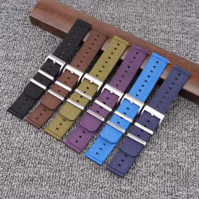 A flat lay of six two-piece nylon watch straps in various colors (black, brown, olive green, purple, light blue, navy blue), each with a silver buckle, fanned out on a grey surface with a wooden block behind.
