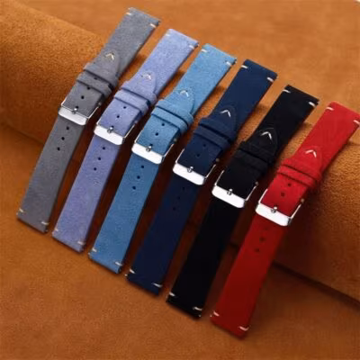 Collection of six vintage suede replacement watch straps arranged diagonally on a brown leather surface. Colors include grey, light blue, mid-blue, navy, black, and red.